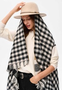 Czarnobiała szachownicowa chusta z frędzlami, noszona na beżowym swetrze, dopełniona jasnobeżowym filcowym kapeluszem z dekoracyjną wstążką.