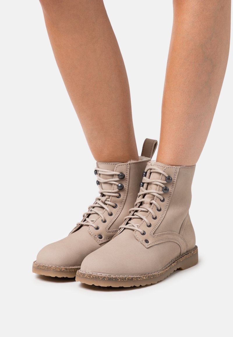 Beige Schnürstiefeletten mit schwarzen Metallösen und Gummisohlen, die auf nackten Beinen vor einem weißen Hintergrund getragen werden.