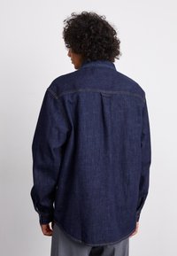 Dark denim shirt with a relaxed fit, long sleeves, button cuffs, and a back yoke detail. Features contrasting stitching and a curved hem.