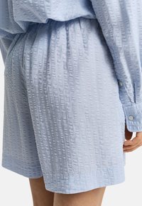 Šviesiai mėlynos šortai, pagaminti iš tiksliai glamžytos medžiagos. Turi elastingą juosmenį ir šonines kišenes. Matomas mygtukais užsegamas rankovės detalė.