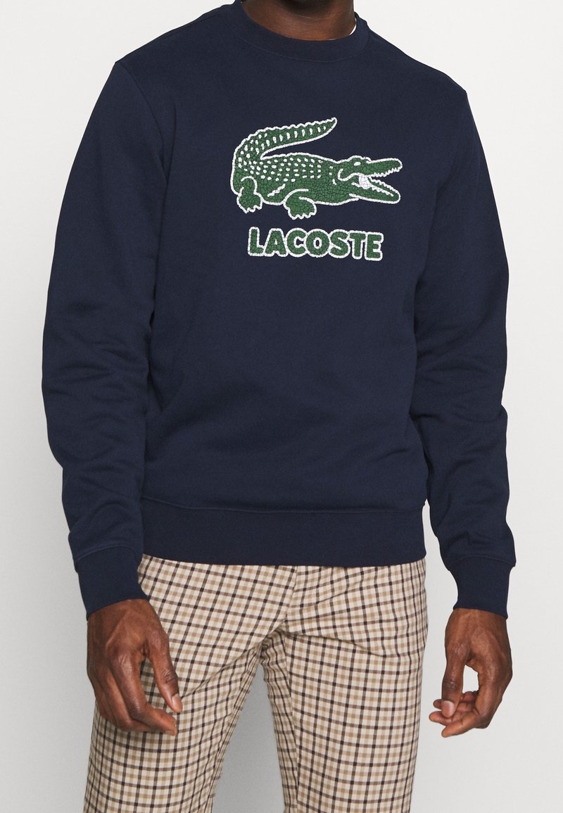 Homme portant un sweat-shirt bleu marine avec logo crocodile vert Lacoste et pantalon beige à carreaux sur fond uni.