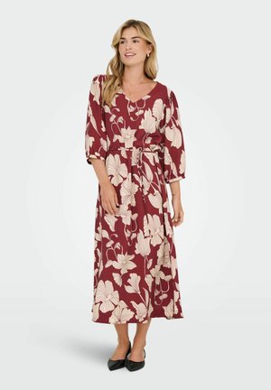 Lång burgundröd klänning med vita blommönster, v-ringad, 3/4 ärmar, åtsittande midja, slät textur och hamn som når anklarna. Kombinerad med svarta klackar.
