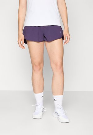 Pantalones cortos deportivos morados con un dobladillo en curva, que presentan un sutil acento de logo. Material ligero combinado con una camiseta blanca que absorbe la humedad y zapatillas blancas.