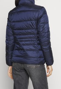 Giacca a piumino blu navy con design trapuntato, colletto alto e polsini elastici. Materiale liscio e lucido con dettagli di cuciture orizzontali.