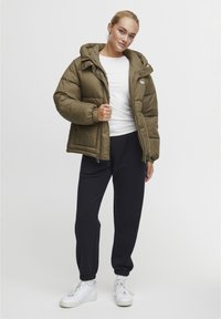Khaki puffer jacket with a hood, elastic cuffs, and zip closure, paired with navy sweatpants and white sneakers, set against a plain background.