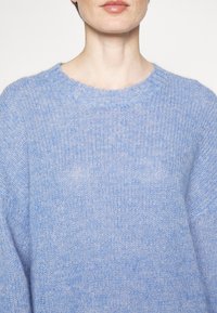 Gros plan d'une personne portant un pull en laine doux bleu avec un col rond, ne montrant que le cou, le menton et le haut du torse.