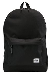 Sac à dos en tissu noir avec poche avant zippée et poignée supérieure, comportant une petite étiquette de marque rectangulaire blanche dans le coin inférieur droit de la poche.