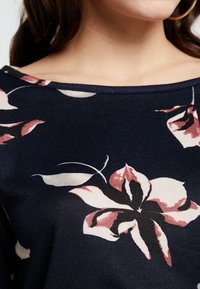 Granatowa bluzka z kwiatowym wzorem w różu i bieli. Posiada szeroki dekolt i fakturowany materiał. Zbliżenie ukazuje szczegóły wzoru.