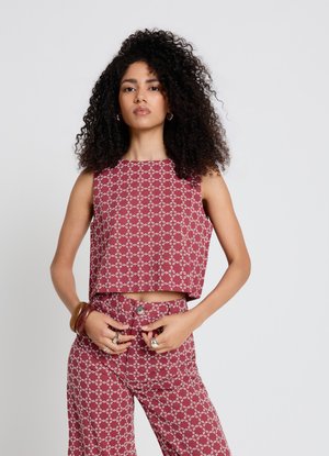Frau mit lockigem Haar, die einen passenden ärmellosen roten Top und Hose mit weißem Kettenglied-Muster trägt, accessoriert mit Ringen und Armreifen.