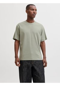 Άνδρας που φοράει ανοιχτό πράσινο T-shirt με στρογγυλή λαιμόκοψη και σκούρο μπλε τζιν, στέκεται μπροστά σε απλό λευκό φόντο, κοιτάζοντας προς τα πλάγια.