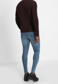 Gebreide kastanjebruine trui, aansluitende blauwe jeans en zwarte sportschoenen. De trui heeft een gestructureerd patroon en een ronde hals.