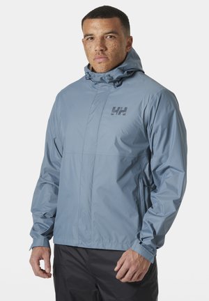 Man met kort haar en sproeten, gekleed in een lichtblauwe regenjas met capuchon en een zwarte broek, staand tegen een lichte achtergrond.