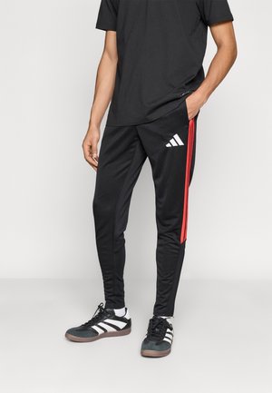 Mężczyzna w czarnych spodniach dresowych Adidas z czerwonymi bocznymi paskami i czarno-białych butach Adidas na jednolitym jasnym tle.