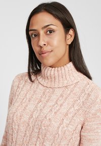 Knitted sweater in light pink with a high ribbed collar. Features cable knit patterns and a textured finish.