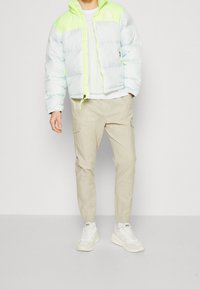 Jasnoniebieska puchowa kurtka z neonowozielonymi akcentami, zamek błyskawiczny i kaptur. Połączona z beżowymi spodniami cargo i białymi trampkami.