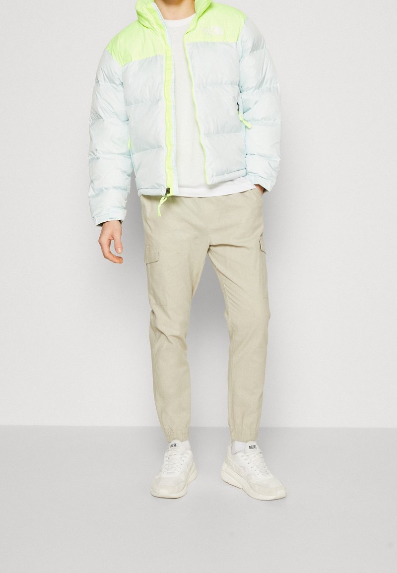 Jasnoniebieska puchowa kurtka z neonowozielonymi akcentami, zamek błyskawiczny i kaptur. Połączona z beżowymi spodniami cargo i białymi trampkami.