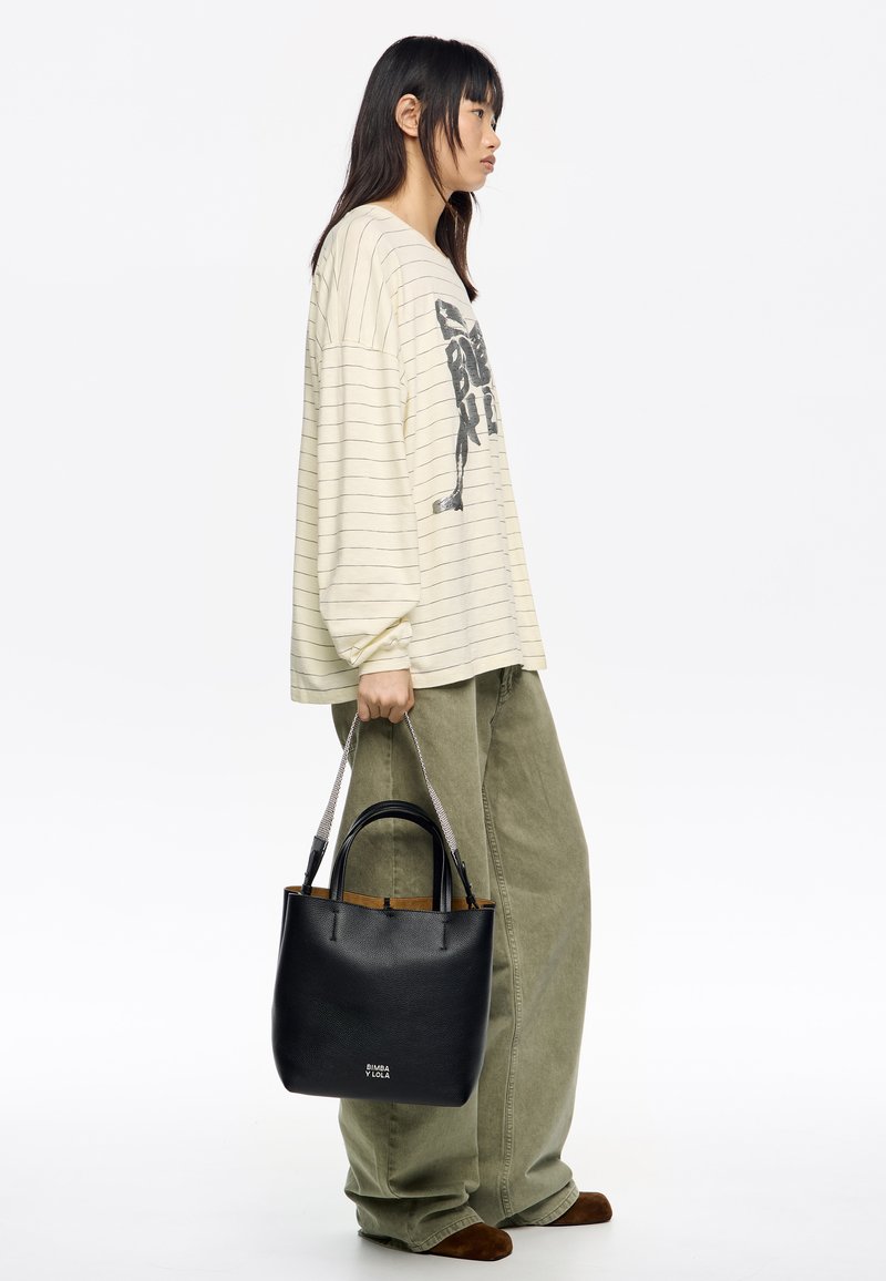 Czarna skórzana torba typu tote z srebrnymi uchwytami w formie łańcucha, z fakturowaną powierzchnią i kontrastowym brązowym wnętrzem, trzymana przez osobę w codziennym stroju.