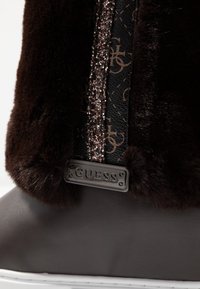 Bota de piel sintética marrón con una banda negra con brillo y un patrón grabado. Incluye una placa con el logo "GUESS" plateada en el lateral.