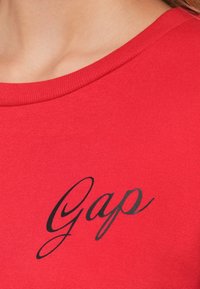 Červená bavlněná mikina s kulatým výstřihem a černým logem "Gap" v písmu na hrudi. Hladká textura, neformální design.