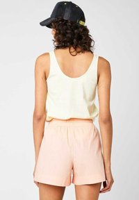 Kobieta z kręconymi włosami, nosząca czarną czapkę, bezrękawnik w kolorze kremowym oraz jasnoróżowe szorty na elastycznym pasie, przedstawiona od tyłu na jednolitym tle.