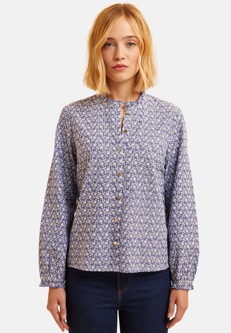 Blusa floreale blu con maniche lunghe, scollatura rotonda e chiusura a bottoni. Presenta un motivo di fiori bianchi e dettagli delle maniche arricciati.
