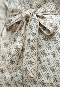 Béžová tkanina s diamantovým vzorom, na ktorom sa opakujú malé logá v hnedej a čiernej farbe. Pásiková stužka siahajúca v páse, lemovaná krémovou farbou.