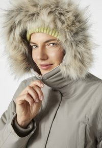 Abrigo de invierno beige con una gran capucha de piel sintética. El cuello está cerrado con cremallera y un gorro de punto verde asoma por debajo de la capucha.