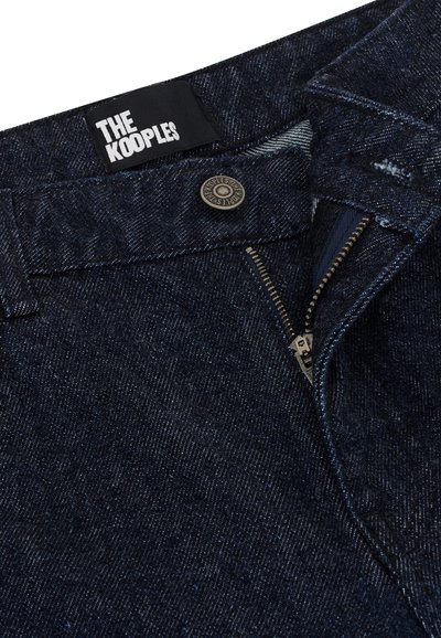 Jeans de denim azul marinho com fecho de zíper, botão em bronze e uma etiqueta com a inscrição "THE KOOPLES." A vista em close destaca a textura e a costura.