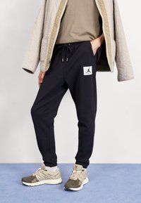 Svarta joggerbyxor med elastisk midja och snörning, med en vit logotyp på lappen. Parat med en ljusbeige jacka och sneakers.