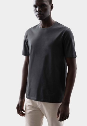 Maglietta a maniche corte di colore grigio scuro in tessuto liscio e leggero; scollo rotondo; taglio aderente. Indossata con pantaloni di colore chiaro.