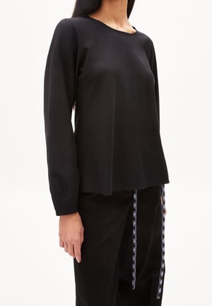 Frau trägt ein schlichtes schwarzes Langarmshirt kombiniert mit schwarzen Hosen, die schwarz-weiße gemusterte Kordeln haben.