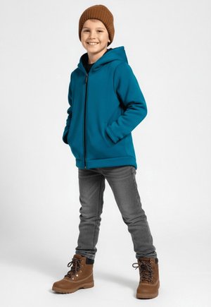 Młody chłopiec uśmiechający się, ubrany w brązową czapkę z dzianiny, niebieskozieloną bluzę zapinaną na zamek, szare dżinsy i brązowe buty sznurowane, stojący z rękami w kieszeniach.