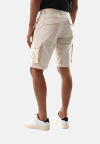 Pantaloni cargo beige con due tasche laterali, tasche posteriori e orli arrotolati. Indossati con sneakers bianche con un accento nero. Tessuto a trama semplice.