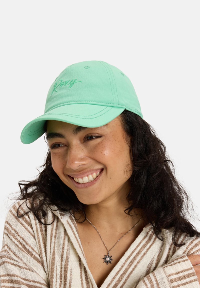 Mint green baseball cap with embroidered "Roxy" logo, curved brim, and eyelets. Worn by a person with curly hair, against a plain background.