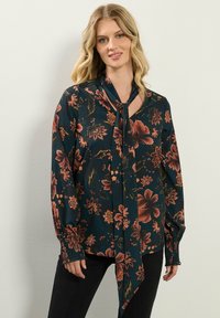 Bloes met bloemenprint in donkergroen met rode en oranje bloemen, lange mouwen, strikdetail bij de hals en gerimpelde boorden. Gladde stof, elegante uitstraling.