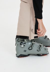 Skischoen in grijs materiaal met een gestructureerd oppervlak en drie gespen. Beigepantalon met rits aan de zoom. Naadloos ontwerp en geventileerd detail.