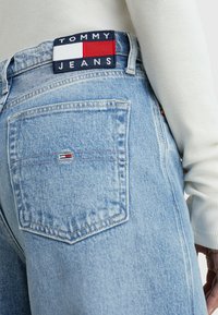 Jeans de mezclilla azul claro con un parche en la parte trasera que presenta "TOMMY JEANS" en colores marino, rojo y blanco. El bolsillo trasero tiene un detalle del logo de la marca.