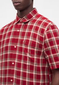 Άνδρας που φοράει κόκκινο καρό πουκάμισο με κοντά μανίκια και τσέπη στο στήθος, εμφανιζόμενος από το λαιμό μέχρι τη μέση του κορμού σε απλό φόντο.