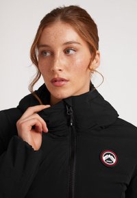 Sort polstret jakke med høj krave, der har en lynlås foran og en rund logo-patch i rød og hvid på brystet. Blød tekstur.