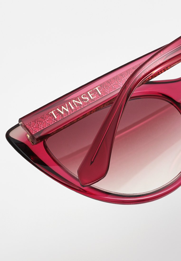 Rote Cat-Eye-Sonnenbrille mit Verlaufsgläsern, ausgestattet mit einem strukturierten Bügel, der das goldene "TWINESET"-Logo trägt. Glatte Flächen und scharfe Winkel.