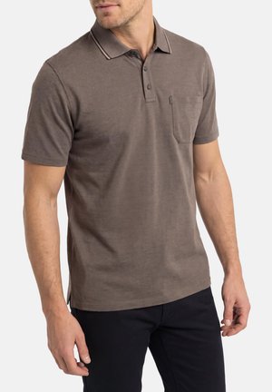 Homme portant un polo marron à manches courtes avec une poche poitrine et un pantalon noir sur fond blanc.