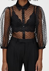 Zwarte transparante blouse met pofmouwen en stippenpatroon, met een knoopsluiting aan de voorkant en een bijpassende zwarte kanten bralette eronder.