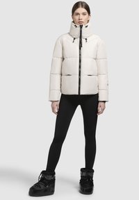 Doudoune gris clair avec un col haut, une fermeture éclair noire et des détails à cordon. Associée à des leggings noirs et des bottes.