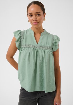Femme avec des taches de rousseur portant des boucles d'oreilles créoles en or et un chemisier vert menthe avec un bord en dentelle et des manches courtes à volants, debout sur fond blanc.