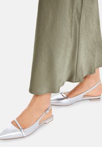 Silberne metallische Slingback-Ballerinas mit spitzem Zehenbereich und minimalen Riemen-Details. Texturierte Oberfläche, flacher Absatz, kombiniert mit einem olivgrünen Kleidungsstück.