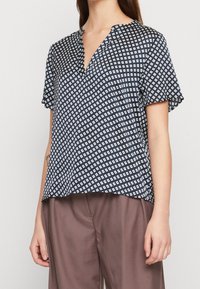 Blusa corta blu navy con motivo a diamante bianco. Presenta un collo a V e una vestibilità rilassata, realizzata in tessuto morbido. Abbinata a pantaloni marroni.