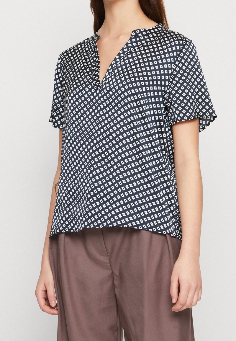 Blusa corta blu navy con motivo a diamante bianco. Presenta un collo a V e una vestibilità rilassata, realizzata in tessuto morbido. Abbinata a pantaloni marroni.