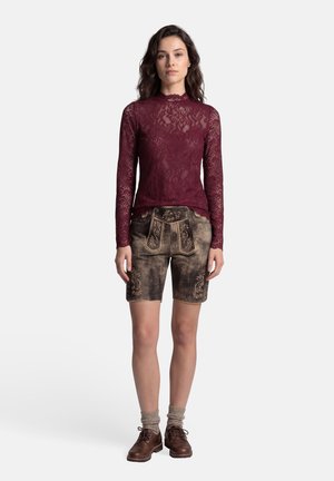 Vrouw met een bordeauxrode kanten top met lange mouwen, bruine leren korte broek met borduursel, beige sokken enbruine veterschoenen, staand tegen een witte achtergrond.