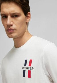 Witte katoenen t-shirt met het "HECHTER" logo in zwart, met verticale blauwe en rode strepen aan de linkerkant. Gladde textuur, ronde halsontwerp.
