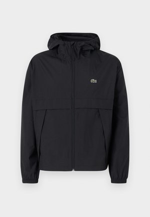 Chaqueta cortaviento negra con cremallera y capucha, puños elásticos, bolsillos laterales y pequeño logo de cocodrilo verde en el pecho.